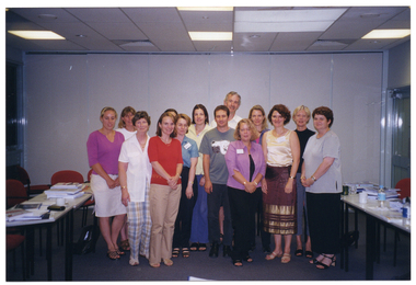 COL photo of a group of people, standing in the middle of a large room. On either side of them, there are several long rectangular tables joined together, with chairs on one side. On the tables, there are several stacks of books and other paperwork, cups and saucers, mugs, and some plastic water bottles. In the background, there is a set of closed bi-fold doors forming a wall behind them.