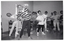 B&W photo of a group of people exercising in a large room. They are standing with their hands on their hips and bending their torsos to one side. Each person has a chair behind them. In the background, there is an open doorway and an "EXIT" sign jutting out from the wall.