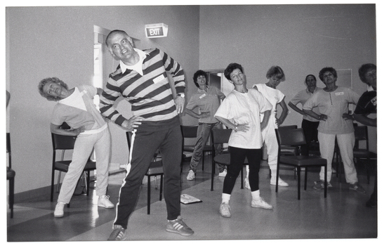 B&W photo of a group of people exercising in a large room. They are standing with their hands on their hips and bending their torsos to one side. Each person has a chair behind them. In the background, there is an open doorway and an "EXIT" sign jutting out from the wall.
