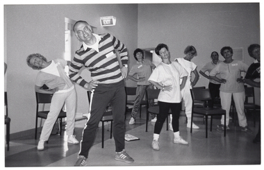 B&W photo of a group of people exercising in a large room. They are standing with their hands on their hips and bending their torsos to one side. Each person has a chair behind them. In the background, there is an open doorway and an "EXIT" sign jutting out from the wall.