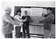 B&W photo of a man and three women standing in front of a large freestanding whiteboard. They are each holding a stretch band and, using both hands, stretching it in front of them at chest level. Affixed to the whiteboard with large bulldog clips is a notice with the text: "TYPES OF EXERCISE 1. Stretching - range of movement -"Coax" - do not bounce! -3-10 reps., 2-4 times per day 2. Strengthening -Minimize stress - "isometrics" -3-4 reps., once a day -Hold each rep. "6 seconds" 3. Endurance -Choose appropriate type -Begin slowly, build progressively". On the right side of the notice, the above text has been grouped together with an arrow-shaped bracket, indicated by two diagonal lines forming an arrow and pointing to the text: "TWO HOUR PAIN RULE". Along the wall behind the whiteboard is a large blackboard, with several cupboards below it. There is some transference of blue ink on the photo image.
