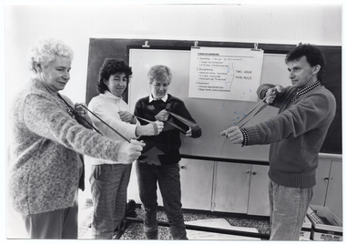 B&W photo of a man and three women standing in front of a large freestanding whiteboard. They are each holding a stretch band and, using both hands, stretching it in front of them at chest level. Affixed to the whiteboard with large bulldog clips is a notice with the text: "TYPES OF EXERCISE 1. Stretching - range of movement -"Coax" - do not bounce! -3-10 reps., 2-4 times per day 2. Strengthening -Minimize stress - "isometrics" -3-4 reps., once a day -Hold each rep. "6 seconds" 3. Endurance -Choose appropriate type -Begin slowly, build progressively". On the right side of the notice, the above text has been grouped together with an arrow-shaped bracket, indicated by two diagonal lines forming an arrow and pointing to the text: "TWO HOUR PAIN RULE". Along the wall behind the whiteboard is a large blackboard, with several cupboards below it. There is some transference of blue ink on the photo image.
