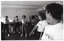 B&W photo of a group of people exercising inside a large room. They are standing and twisting their torsos to one side from the waist. Some people are wearing t-shirts with the "Joint Action" logo, some are wearing t-shirts with a different logo (indistinguishable), while others are wearing contemporary clothing. Behind them, there is a large billiard table with a few billiard balls on top. In the background, against the wall, there is a cue rack with three cues, and above it, a clock on the wall.