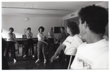 B&W photo of a group of people exercising inside a large room. They are standing and twisting their torsos to one side from the waist. Some people are wearing t-shirts with the "Joint Action" logo, some are wearing t-shirts with a different logo (indistinguishable), while others are wearing contemporary clothing. Behind them, there is a large billiard table with a few billiard balls on top. In the background, against the wall, there is a cue rack with three cues, and above it, a clock on the wall.