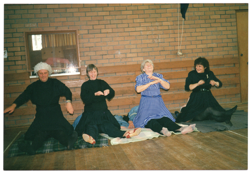 COL photo of four people lying face-up on some blankets on the wooden floor, with only their legs showing. Meanwhile, four other people are sitting across them with their knees on the blankets. Some of the people kneeling are laughing. In the background, there is a brick wall with a window, through which a storage room with some cardboard boxes is evident.