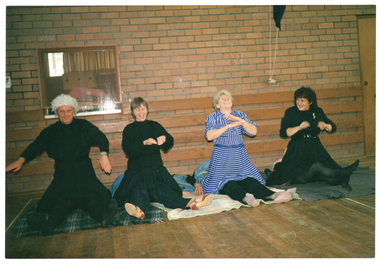 COL photo of four people lying face-up on some blankets on the wooden floor, with only their legs showing. Meanwhile, four other people are sitting across them with their knees on the blankets. Some of the people kneeling are laughing. In the background, there is a brick wall with a window, through which a storage room with some cardboard boxes is evident.