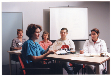 COL photo of a small group of people sitting around a rectangular table indoors. One woman is holding up a circular piece of cardboard or plastic. It is titled, "The Hip", and contains some text and a large illustration of a hip joint and pelvis. There is another similar disc on the table. The other four people are all looking at the woman with the disc. They each have several books and folders on the table in front of them. Behind them, there is a freestanding slide projector screen near the wall.