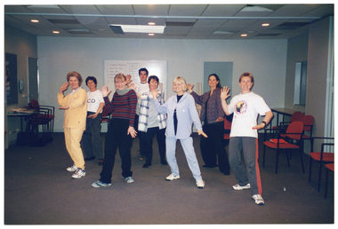 COL photo of a large group of people standing with one arm up (palm outward), one arm down (palm facing the floor), and their feet apart with one leg slightly forward. They are in a large room, with a few tables and several chairs around the periphery of the room. There is a large whiteboard affixed to the wall behind them.