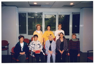 COL photo of eight women in a large room. Five of the women are sitting on a row of chairs, with the remaining three women standing behind them. One of the women in the front row is holding a red folder in her lap. In the background, there are some large windows with thin venetian blinds and a view to some large ferns outside. To the right of the frame, there is a set of tall bi-fold doors in the open position. There are a few empty chairs around the room.