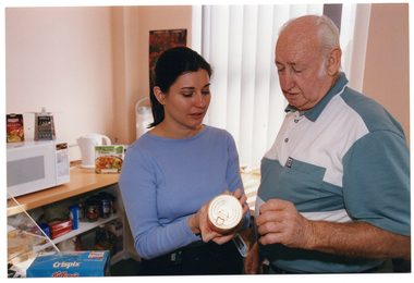 COL photo of a woman and a man standing next to one another in a kitchen. The woman is holding a can of food in one hand and pointing out the information on the label to the man. On a table or bench in front of them is a box of Kellog's Crispix cereal. Against the wall behind them are some shelves stocked with packaged food/pantry items. On the bench above the shelves, there is a microwave oven, an electric kettle, and an urn (partially obscured). On top of the microwave oven, there is a metal grater and a small carton of packaged food, with a slightly bigger carton packaged food on the bench in front of the kettle. There is a poster on the wall behind the urn, which sits beside a window with vertical blinds.