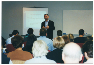 COL photo of a man standing in front of a roomful of people who are seated. Behind him, there is a slide projector screen with a slide being projected titled, "Self Management Does it make a difference?" Below the title is the text: "AFV Centre for Rheumatic Diseases The University of Melbourne Royal Melbourne Hospital". On the wall behind him, there is a large whiteboard. In the foreground, there are several people seated, who have been Photographed from behind.
