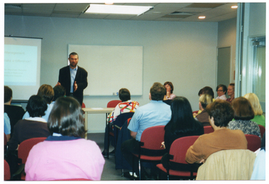 COL photo of a man standing in front of a roomful of people who are seated. Behind him, there is a slide projector screen with a slide being projected titled, "Self Management Does it make a difference?" Below the title is the text: "AFV Centre for Rheumatic Diseases The University of Melbourne Royal Melbourne Hospital". On the wall behind him, there is a large whiteboard with a table beneath it. In the foreground, there are several people seated, who have been Photographed from behind. This photo has been taken at a slight angle to the left of the frame.