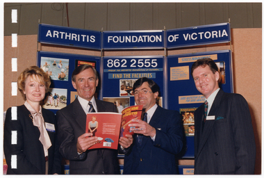 COL photo of four people standing next to one another in front of a three-panelled display board. The two people in the middle are holding an open book titled, "Exercise beats Arthritis". At the top of the display board is the text, "ARTHRITIS FOUNDATION OF VICTORIA", with the telephone number "862 2555" below that.