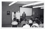 B&W photo of three people in front of a seated audience. A woman and a man are standing on either side of a freestanding whiteboard, while another woman is sitting at a large rectangular table. Behind them, there is a large projector screen affixed to the brick wall. There are two posters on the wall, one of which contains the text "Arthritis affects one in five Australians". There are also two handwritten signs on the wall, one of which contains the title, "BINGO".