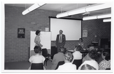 B&W photo of three people in front of a seated audience. A woman and a man are standing on either side of a freestanding whiteboard, while another woman is sitting at a large rectangular table. Behind them, there is a large projector screen affixed to the brick wall. There are two posters on the wall, one of which contains the text "Arthritis affects one in five Australians". There are also two handwritten signs on the wall, one of which contains the title, "BINGO".