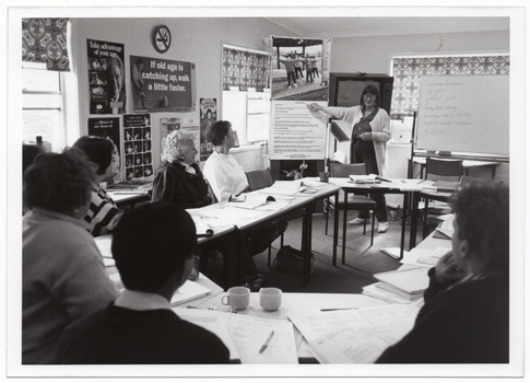 B&W photo of a woman standing at one end of a large room, facing a group of people who are seated at tables. She is pointing to a large piece of paper with text, above which there is a large poster of a group of people exercising. Beside her, is a large freestanding whiteboard, with some handwritten text on it. Behind her, there is a television set on a tall stand. The tables have been arranged in an oval configuration. On top of the tables, there are books and folders of printed material in front of every person, including the woman standing, and a few cups or mugs. There are several posters on the walls, containing text such as, "Take advantage of your age", "If old age is catching up, walk a little faster", "....Move it or lose it.", and "Table tennis keeps you moving". Around the room, there are large windows with holland blinds with a bold pattern.