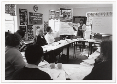 B&W photo of a woman standing at one end of a large room, facing a group of people who are seated at tables. She is pointing to a large piece of paper with text, above which there is a large poster of a group of people exercising. Beside her, is a large freestanding whiteboard, with some handwritten text on it. Behind her, there is a television set on a tall stand. The tables have been arranged in an oval configuration. On top of the tables, there are books and folders of printed material in front of every person, including the woman standing, and a few cups or mugs. There are several posters on the walls, containing text such as, "Take advantage of your age", "If old age is catching up, walk a little faster", "....Move it or lose it.", and "Table tennis keeps you moving". Around the room, there are large windows with holland blinds with a bold pattern.