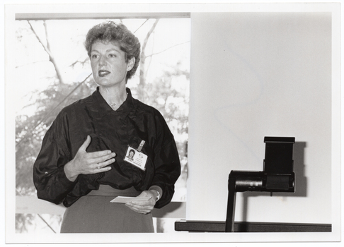B&W medium close up photo of a woman standing in front of a window, speaking. Beside her, there is an overhead slide projector. There is a view of some trees through the window behind her. One side of the window has the blind drawn down.