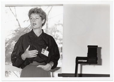 B&W medium close up photo of a woman standing in front of a window, speaking. Beside her, there is an overhead slide projector. There is a view of some trees through the window behind her. One side of the window has the blind drawn down.