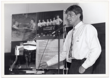 B&W medium close up photo of a man speaking into a microphone on a stand. Beside him, there is an overhead slide projector. Behind him, there is a display board containing three large posters and a section with some printed text. The posters contain images pertaning to water exercise, walking, and a portrait of a group of people standing.