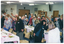 COL photo of a group of people doing some gentle stretching exercises. They are standing behind their chairs (situated at long rectangular tables), and sliding one arm down one side of their bodies. The tables have been set with disposable plates and colourful serviettes, and decorated with ferns and flowers. In the background, an arrangement of dried flowers in a vase sits on a tall stand or cupboard. There are some pictures and a fan affixed to the wall. On the adjoining wall, there is a servery which is closed with a wooden panelled screen. On the top left hand side of the frame around the servery, there is a small sign containing the text, "COFFEE". On another wall, there is a bookcase or dresser with various items, such as vases and framed items, on the shelves. On the wall next to it is a wall phone and a small noticeboard. Above the noticeboard, there is a long rectangular speaker affixed to the wall. There are several ceiling fans suspended from the ceiling.