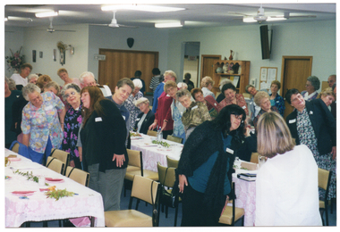 COL photo of a group of people doing some gentle stretching exercises. They are standing behind their chairs (situated at long rectangular tables), and sliding one arm down one side of their bodies. The tables have been set with disposable plates and colourful serviettes, and decorated with ferns and flowers. In the background, an arrangement of dried flowers in a vase sits on a tall stand or cupboard. There are some pictures and a fan affixed to the wall. On the adjoining wall, there is a servery which is closed with a wooden panelled screen. On the top left hand side of the frame around the servery, there is a small sign containing the text, "COFFEE". On another wall, there is a bookcase or dresser with various items, such as vases and framed items, on the shelves. On the wall next to it is a wall phone and a small noticeboard. Above the noticeboard, there is a long rectangular speaker affixed to the wall. There are several ceiling fans suspended from the ceiling.