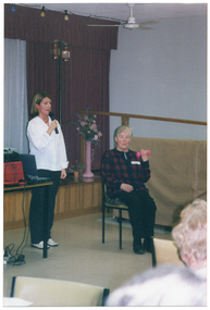 COL photo of a woman is standing beside a table, speaking into a cordless handheld microphone. Beside her, another woman is sitting on a chair: with one arm bent at the elbow, she is holding up a small pink gym hand weight. There are various items piled onto the table. Above the pile is a slide projector with its cord dangling down. The cord is plugged into a power board under the table. Behind them is a stage with long red curtains. At the front of the stage, there is a tall pink vase containing an arrangement of flowers. There is a string of coloured lights hanging from the ceiling in front of the stage, and a ceiling fan.