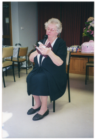 COL photo of a woman sitting on a chair. She is speaking into a cordless handheld microphone while reading from an open booklet. Behind her, there is a stage with the stage curtains closed. On the edge of the stage, there is a large floral arrangement and some gift wrapped items. On the left of the frame, there is a long rectangular table with some chairs. Between the table and the stage, there is a wire shopping trolley leaning against the wall.