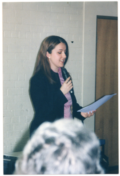 COL photo of a woman speaking into a hand-held microphone while she reads from a sheet of paper. A grey-haired woman's head is evident in the foreground.