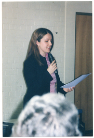 COL photo of a woman speaking into a hand-held microphone while she reads from a sheet of paper. A grey-haired woman's head is evident in the foreground.