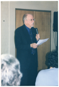 COL photo of a man speaking into a hand-held microphone while holding a piece of paper in his other hand. There is a double wooden door behind him. Next to the door, there is a speaker on a stand (partially obscured). In the foreground, two people have been Photographed from behind.