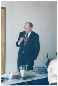 COL photo of a man speaking into a hand-held microphone. There is a large rectangular table in front of him. On the table, there is a slide projector, a wooden tray with a jug of water, two glasses of water, some pieces of paper, and an electrical cable.