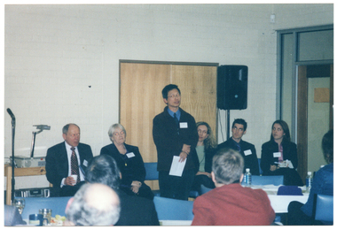 COL photo of a man standing in front of a row of chairs, at which five others are seated. Behind them, there is a slide projector on one side and a speaker on a tall stand on the other side. Seated at tables in the foreground, some members of the audience have been Photographed from behind.