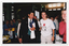 COL photo of a man and two women at a crowded indoor event. The man is wearing a white shirt, black suit, and green tie. He is holding a wrapped gift, an envelope, and a white t-shirt in one hand. One of the women is wearing a t-shirt onto which the w