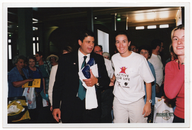 COL photo of a man and two women at a crowded indoor event. The man is wearing a white shirt, black suit, and green tie. He is holding a wrapped gift, an envelope, and a white t-shirt in one hand. One of the women is wearing a t-shirt onto which the w