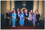 COL photo of a group of people posing for a photo. Some are wearing blue sashes with gold text across their torsos. Behind the group, there is a dais with blue carpet, on which sits a blue upholstered throne with a gold frame. Behind the dais, there is a blue velvet curtain upon which hangs a plaque depicting a crown and the text "E II R".