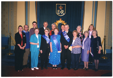 COL photo of a group of people posing for a photo. Some are wearing blue sashes with gold text across their torsos. Behind the group, there is a dais with blue carpet, on which sits a blue upholstered throne with a gold frame. Behind the dais, there is a blue velvet curtain upon which hangs a plaque depicting a crown and the text "E II R".