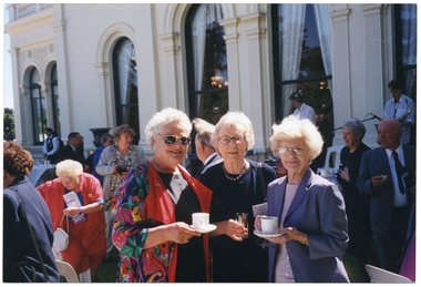 COL photo of three women posing for a photo with a crowd behind them, outside a large building. The woman in the middle is holding a glass, while the women on either side of her are each holding a cup and saucer. There are several people in the background, including two musicians on a small raised stage.
