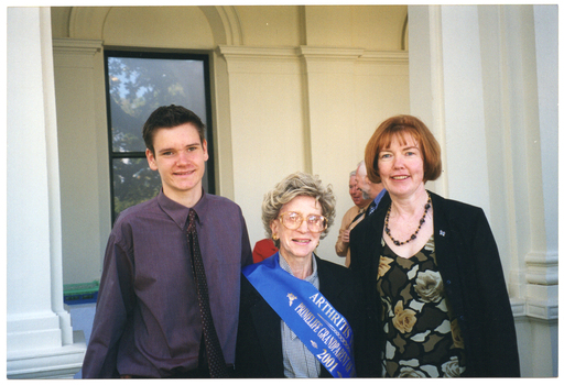 COL photo of a teenage boy, an elderly woman, and a middle-aged woman posing for a photo outside a large building. A few other people are standing in the background.