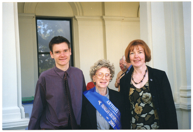 COL photo of a teenage boy, an elderly woman, and a middle-aged woman posing for a photo outside a large building. A few other people are standing in the background.