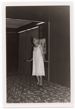 B&W photo of a woman modelling a sleeveless dress, in a plain pale coloured fabric, on a small stage. There is a framed picture on the wall behind her, and some stage curtains behind one section of the stage area. There is a bold patterned carpet on the stage floor.