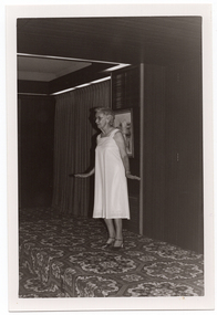 B&W photo of a woman modelling a sleeveless dress, in a plain pale coloured fabric, on a small stage. There is a framed picture on the wall behind her, and some stage curtains behind one section of the stage area. There is a bold patterned carpet on the stage floor.