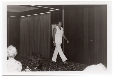 B&W photo of a woman walking across a small stage, modelling a sleeveless tunic and trouser ensemble in a plain pale coloured fabric. There is a framed picture on the wall behind her, and some stage curtains behind one section of the stage area. There is a bold patterned carpet on the stage floor. In the foreground, the heads of a few people from the audience have been Photographed from behind.