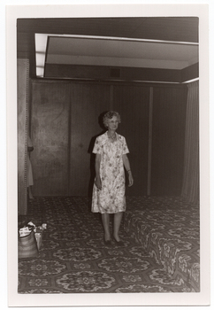B&W photo of a woman modelling a short-sleeved, light-coloured patterned dress on the carpeted floor below a low stage. On the side of the floor and stage, there is a wood-panelled wall and at the back of the stage, a curtain (partially obscurred). There is a bold patterned carpet on the stage and floor. On the floor, to the left of the frame, there is a carry bag and small plastic crate filled with miscellaneous items.