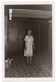 B&W photo of a woman modelling a short-sleeved, light-coloured patterned dress on the carpeted floor below a low stage. On the side of the floor and stage, there is a wood-panelled wall and at the back of the stage, a curtain (partially obscurred). There is a bold patterned carpet on the stage and floor. On the floor, to the left of the frame, there is a carry bag and small plastic crate filled with miscellaneous items.