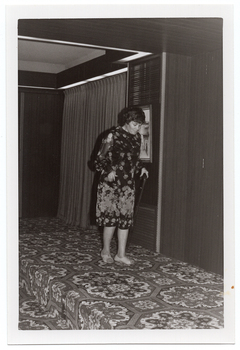 B&W photo of a woman modelling a long-sleeved dark floral dress on a small stage. She is using a walking stick in one hand, and looking down slightly. There is a framed picture on the wall behind her, and some stage curtains behind one section of the stage area. There is a bold patterned carpet on the stage floor.