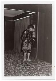 B&W photo of a woman modelling a long-sleeved dark floral dress on a small stage. She is using a walking stick in one hand, and looking down slightly. There is a framed picture on the wall behind her, and some stage curtains behind one section of the stage area. There is a bold patterned carpet on the stage floor.