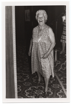 B&W photo of a woman, smiling broadly, modelling a sleeveless multi-coloured floral dress in a light colour. She is on the carpeted floor below a low stage. To the right of the frame, there is a woman (partially obscurred) on the stage, speaking into a microphone on a stand. There is a small shiny wastepaper bin at her feet. In the foreground, to the left of the frame, there is a wooden partition with a decorative edge. There is a bold patterned carpet on the stage and floor.