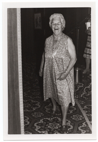 B&W photo of a woman, smiling broadly, modelling a sleeveless multi-coloured floral dress in a light colour. She is on the carpeted floor below a low stage. To the right of the frame, there is a woman (partially obscurred) on the stage, speaking into a microphone on a stand. There is a small shiny wastepaper bin at her feet. In the foreground, to the left of the frame, there is a wooden partition with a decorative edge. There is a bold patterned carpet on the stage and floor.