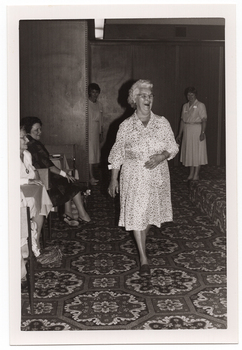 B&W photo of a woman, smiling broadly, modelling a three-quarter sleeve spotted dress in a light colour. She is walking across the bold-patterned carpeted floor below a low stage (which is covered in the same carpet). In the background, there are two other women - one is standing partly behind a wooden partition and the other is standing on the floor beside the low stage. A few members of the seated audience are evident to the left of the frame. There are two or more bags on the floor, at their feet.